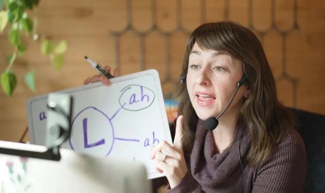 Heather mid-session with a headset, holding a whiteboard showing the letter L and 'ah' sounds