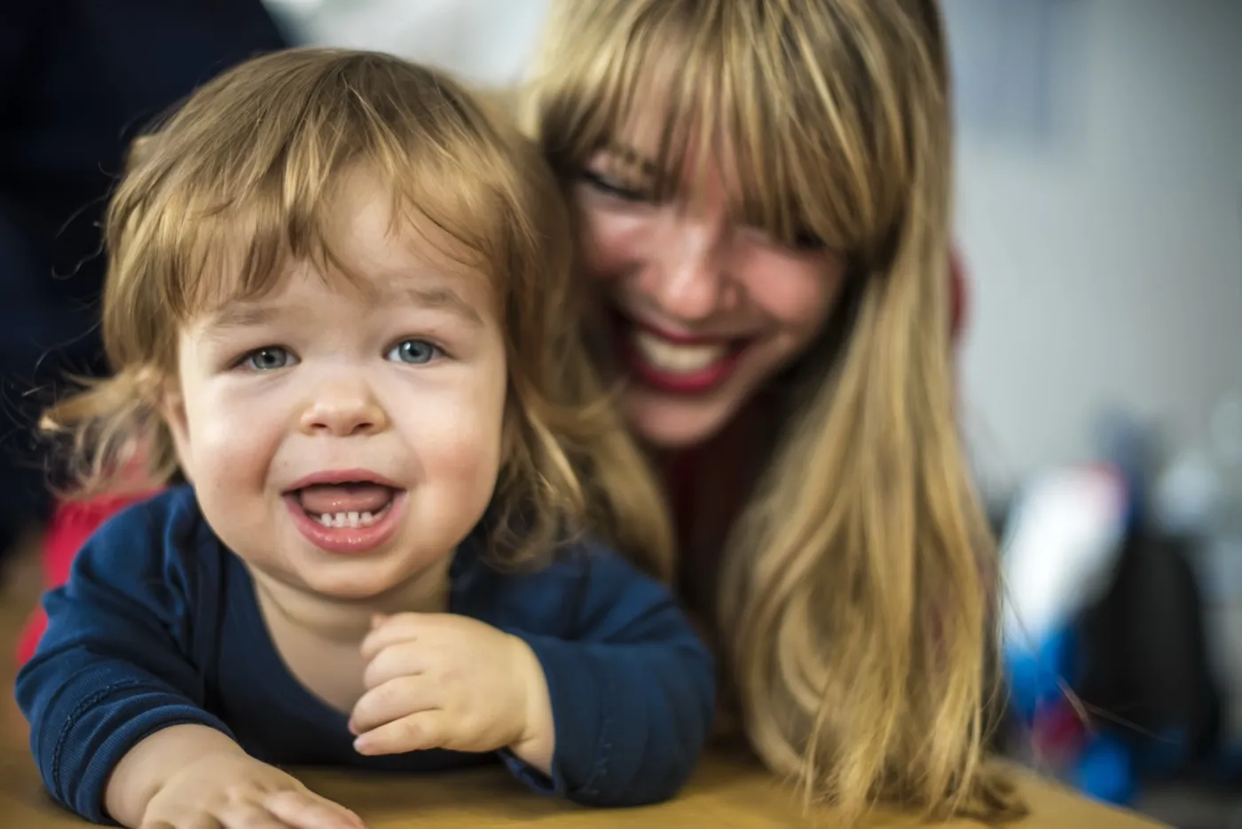 A laughing toddler leaning on a table with a delighted parent close behind